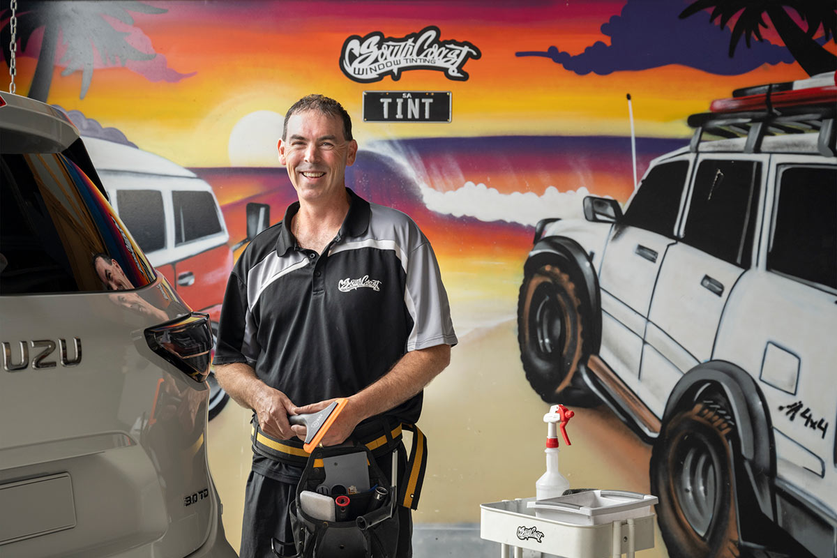 A man smiles at the camera. He has short brown hair and is wearing his uniform and toolbelt. He stands in front of a wall that has a colourful mural of a wave, palm trees and a 4x4 and VW van. The mural reads South Coast Window Tinting.