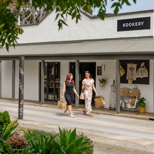 Two women walk down the street smiling and talking to each other. They pass a shop called Kookery displaying clothing and gift items outside.