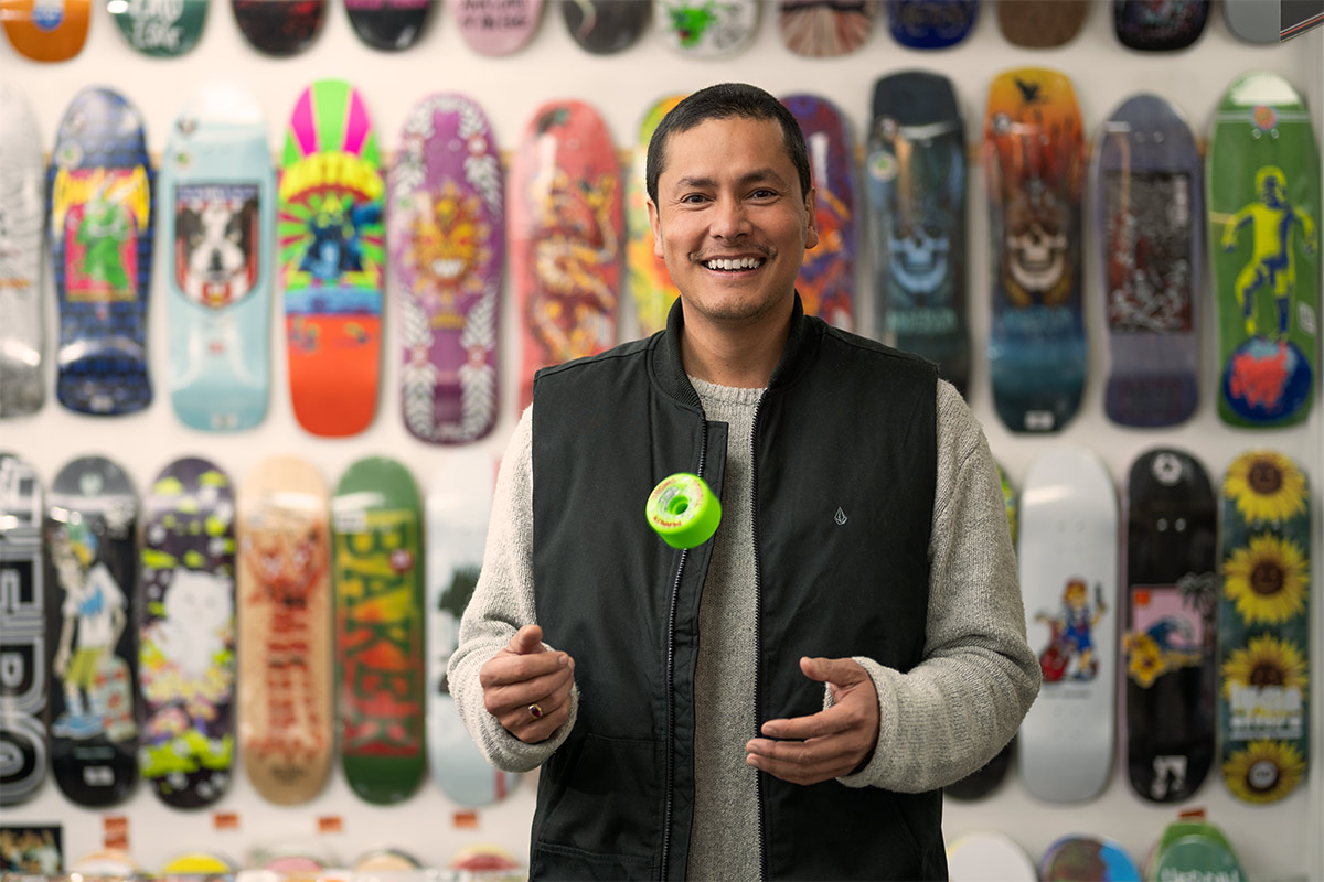 Man throws a skateboard wheel in the air while smiling at camera. Skateboards line the wall behind him.